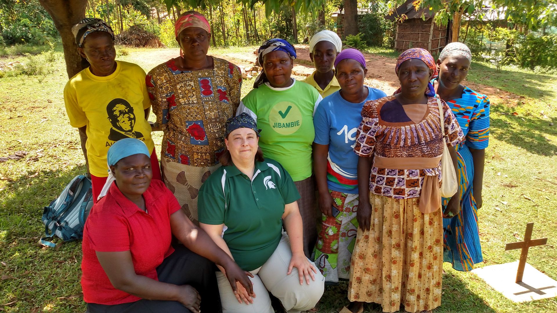 A group of nine women pose together for the camera outdoors. The eight Kenyan women are wearing a variety of colorful clothing and headwraps, with Lisa Tiemann on the left wearing a green Spartan shirt, holding hands with a woman in a red shirt.