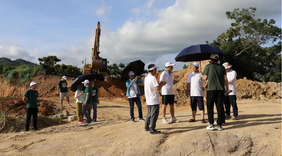 Researchers stand on construction site holding umbrellas to block the hot sun. Mountains and blue sky appear behind them.