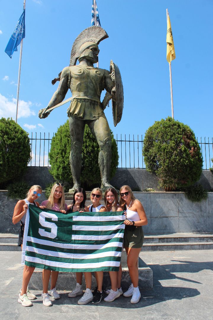 Group of MSU students holding MSU flag in front of a Spartan statue in Greece