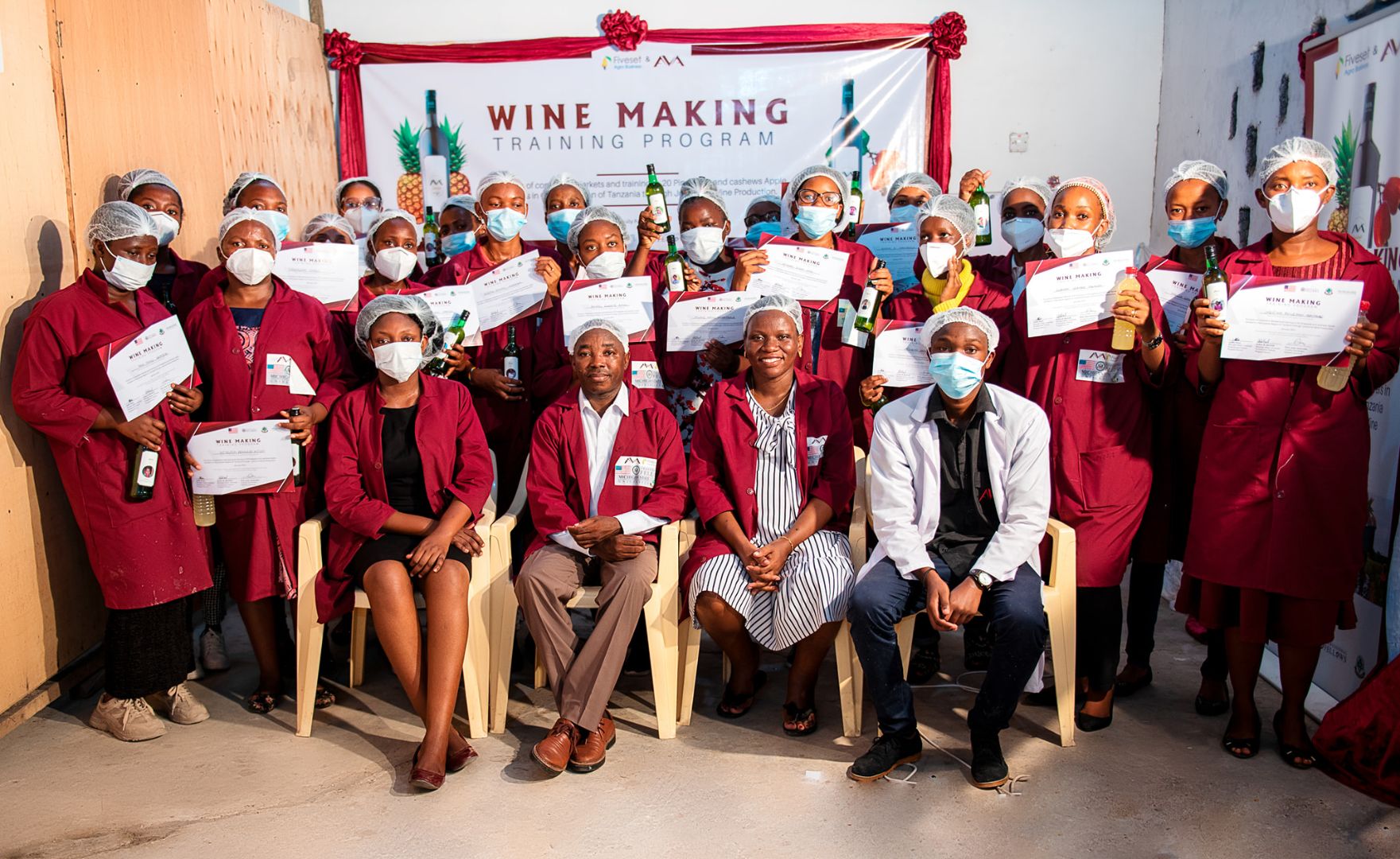 A group of wine making training program participants sit together in red gowns and protective gear holding certificates