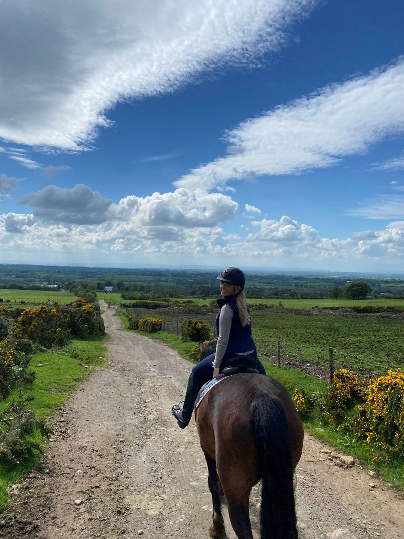 Cora riding a horse on a dirt trail among green pasture in Ireland