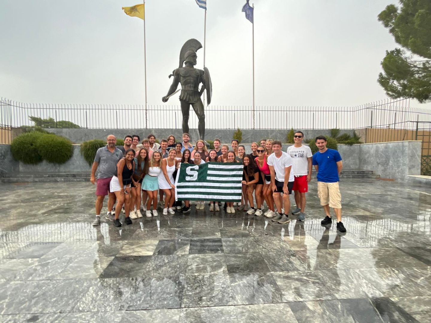 Group of MSU students holding a Spartan flag in front of a Spartan statue in Greece