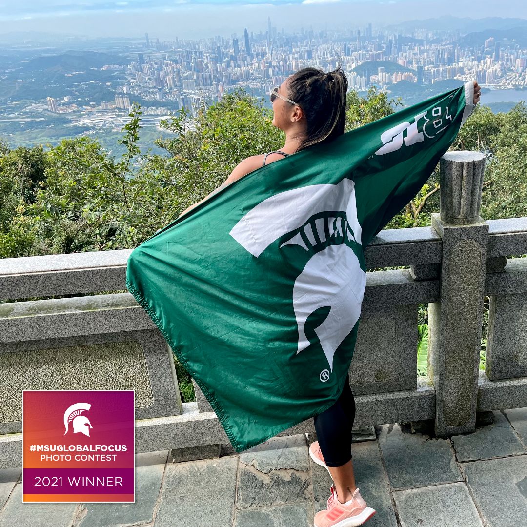 Person with back to the camera holding an outstretched green and white Spartan flag, overlooking a cityscape. Text box with white Spartan helmet "#MSUGLOBALFOCUS Photo Contest 2021 Winner"