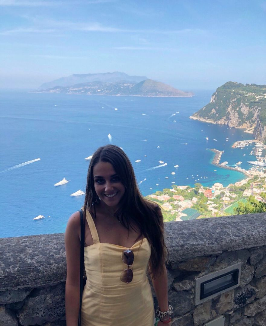 Kierstyn standing by stone wall with sea behind her in Italy