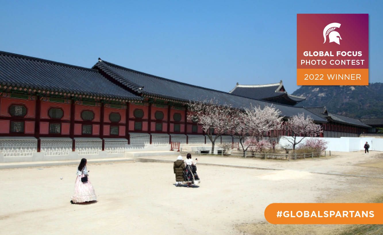 A landscape featuring long building with a tiled roof and a group of blossoming trees. People walk across a dirt plaza in the foreground.