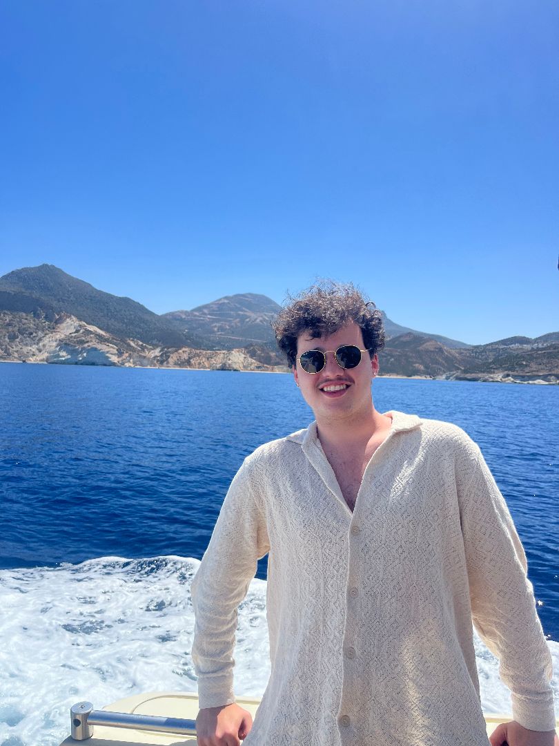 Brennan wearing a white shirt and sunglasses standing by the sea in Greece