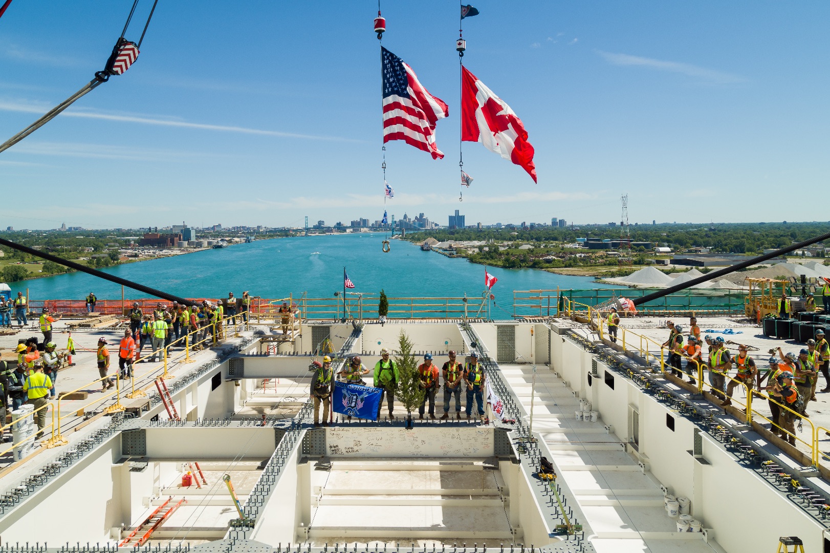 Workers standing on bridge construction site overlooking water with American and Canadian flags flying overhead