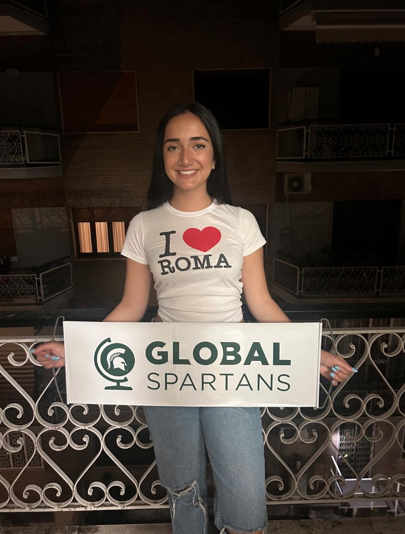 Sasra standing on a balcony at night wearing an I love Roma shirt and holding a Global Spartans banner