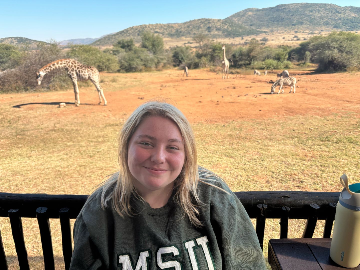 Alexandra wearing an MSU sweatshirt sitting on a balcony with giraffes in the background