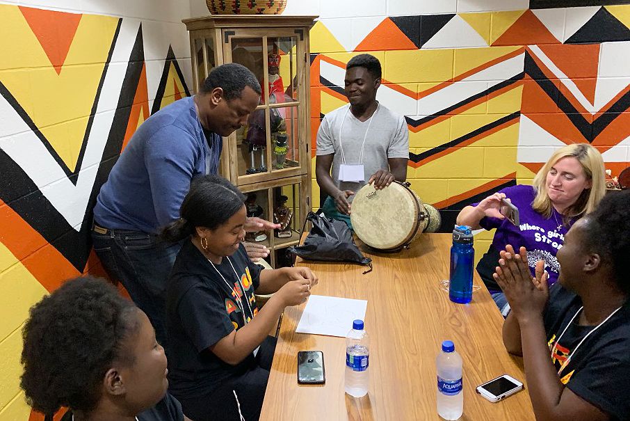 Male standing playing the drum while others gathered at table watch and smile