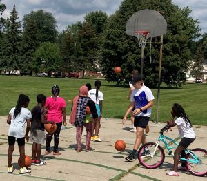 Young children shoot basketballs at a net as MSU students stand by to help rebound