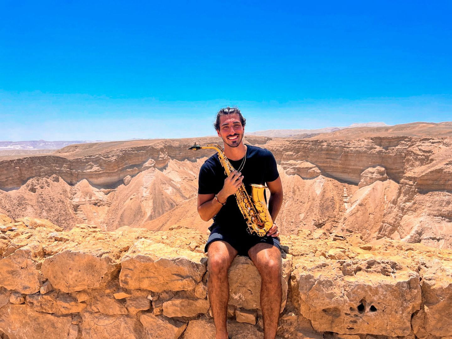 Luke holding his saxophone sitting on a stone wall in the desert