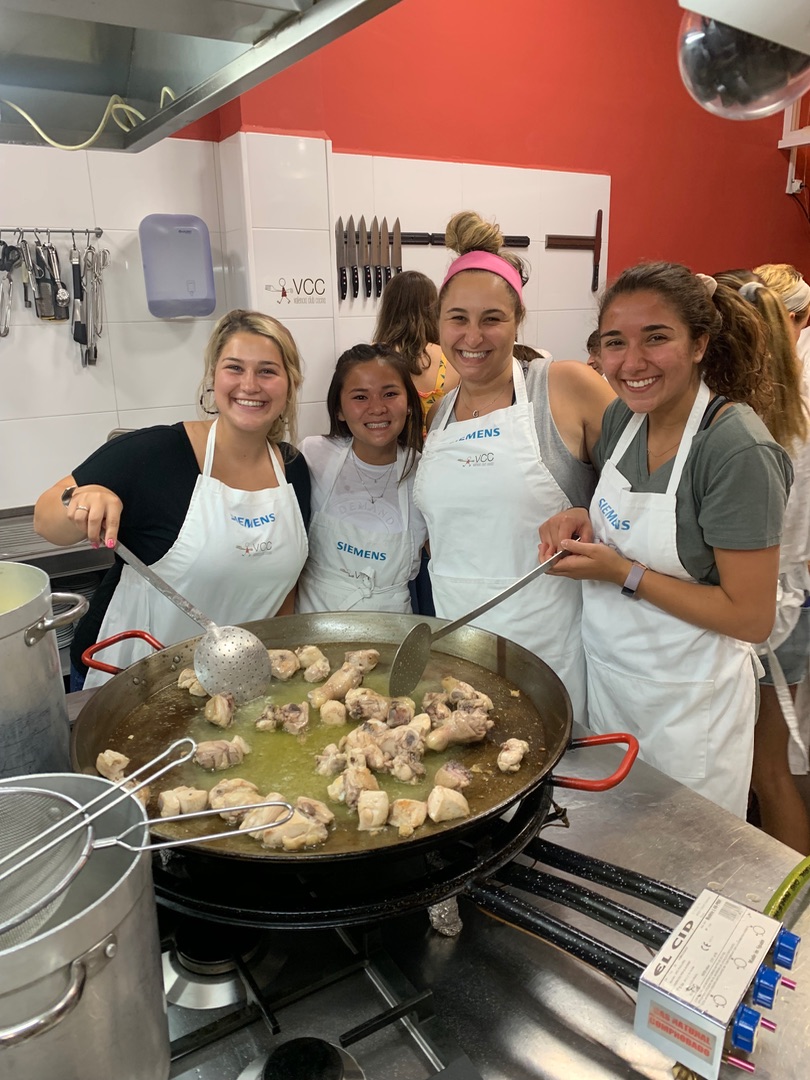 A group of students in aprons posing with a large pot of paella