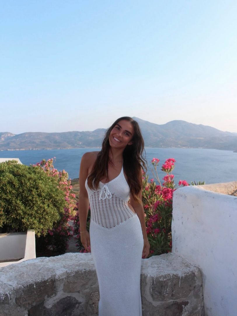 Mya wearing a long white dress standing on a balcony overlooking the sea
