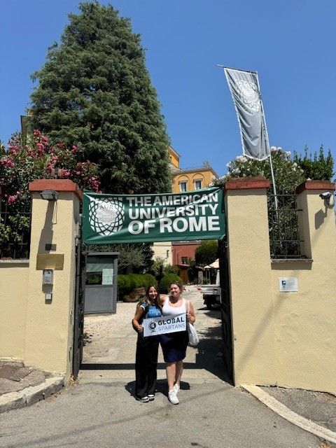 Natalie and friend holding a Global Spartans banner underneath the American University of Rome entrance