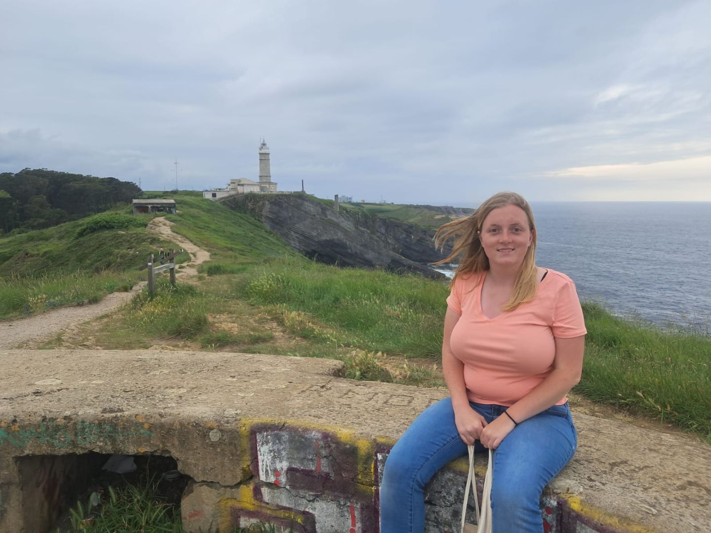 Laurel wearing a pink shirt and jeans sitting on a stone wall by the sea in Spain