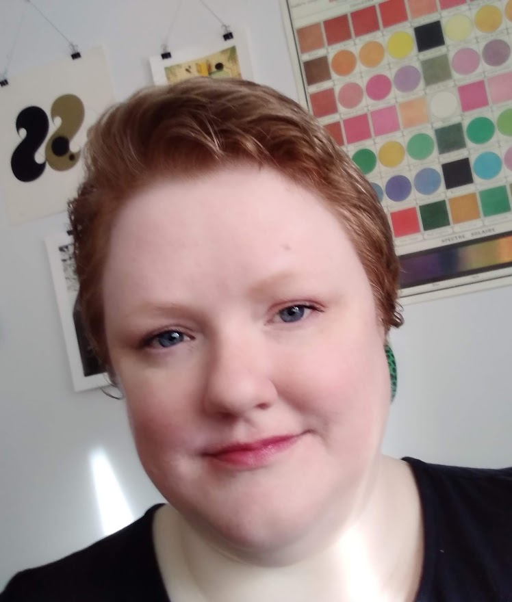 Close up headshot of a smiling woman against a colorful background; she has short red hair and is wearing a black shirt.