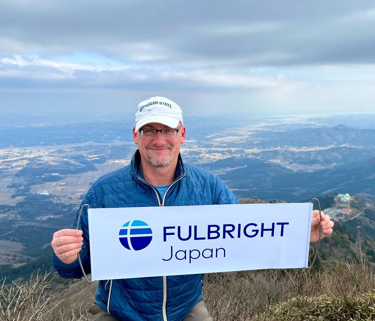 A man in a white Michigan State ball cap, glasses, blue jacket stands on top of a mountain with an expansive landscape below. He holds a rectangular white banner with “Fulbright Japan” printed on it. 