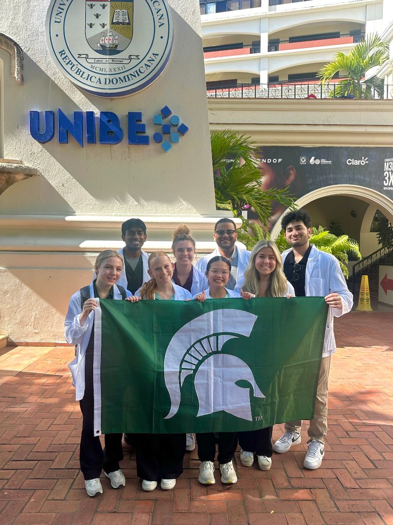 Kaitlynn and friends holding a Spartan flag in front of a building that says UNIBE