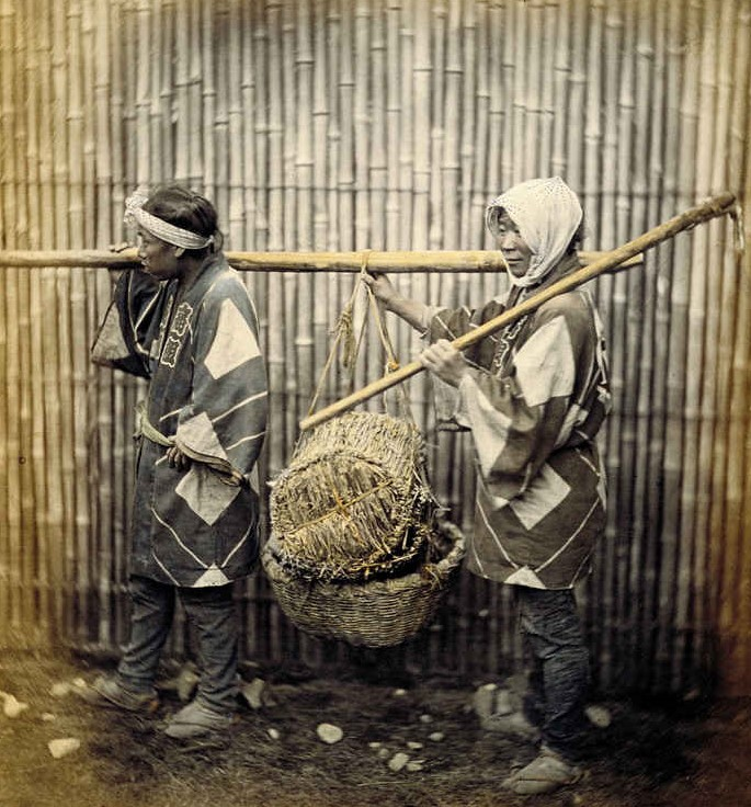an sepia photo of two people in traditional Japanese dress carrying a basket on a long stick