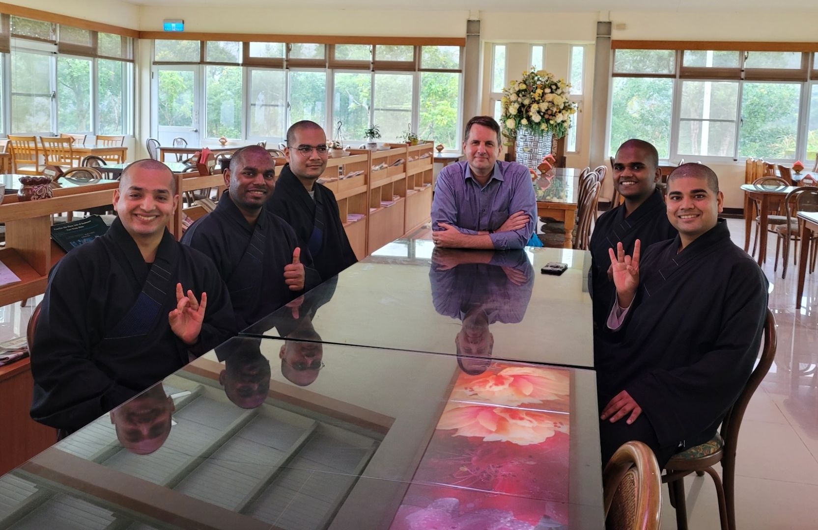 A group of six men sit at a long, reflective table. At left are three men dressed in black Buddhist attire, smiling and gesturing at the camera, a man in a blue shirt in the center crosses his arms, and two men also dressed in black smile and gesture at the camera. A row of windows is in the background.