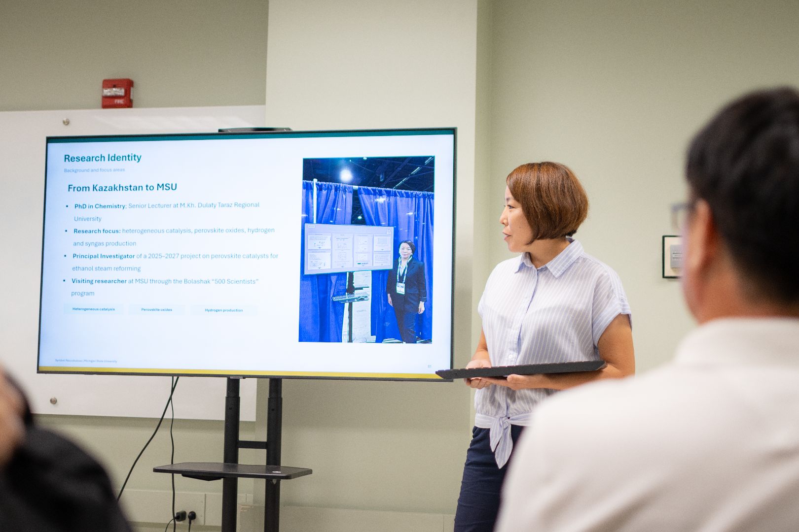 Symbat Naurzkulova in front of a presentation screen and holding a keyboard, speaking to the audience. 