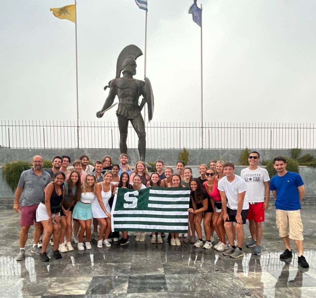 Group of students holding a Spartan flag in front of a Spartan statue in Greece