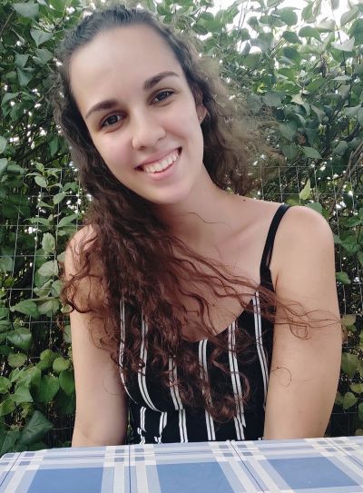 A woman with wavy brown hair and wearing a black and white striped top smiles for the camera in front of a green bush.