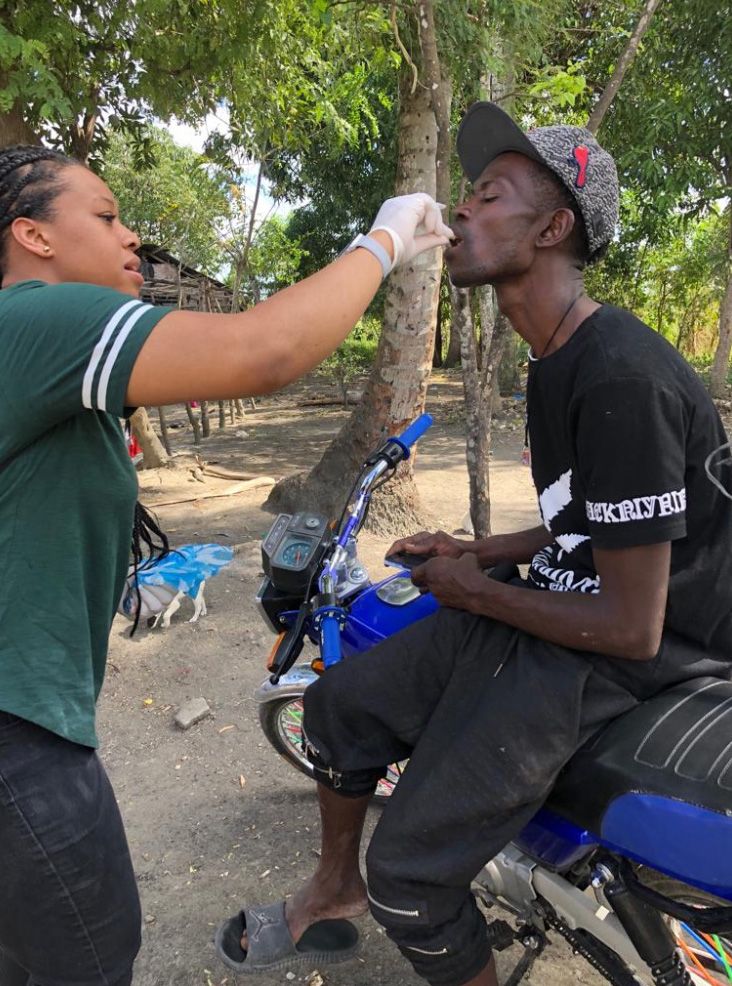 A black woman wearing a green shirt and latex gloves helps a man sitting on a motorbike.