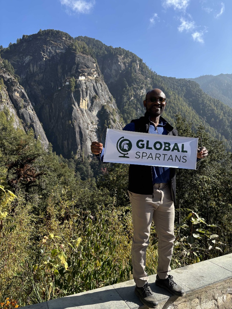 A man poses in front of a mountain while holding a sign that reads Global Spartans