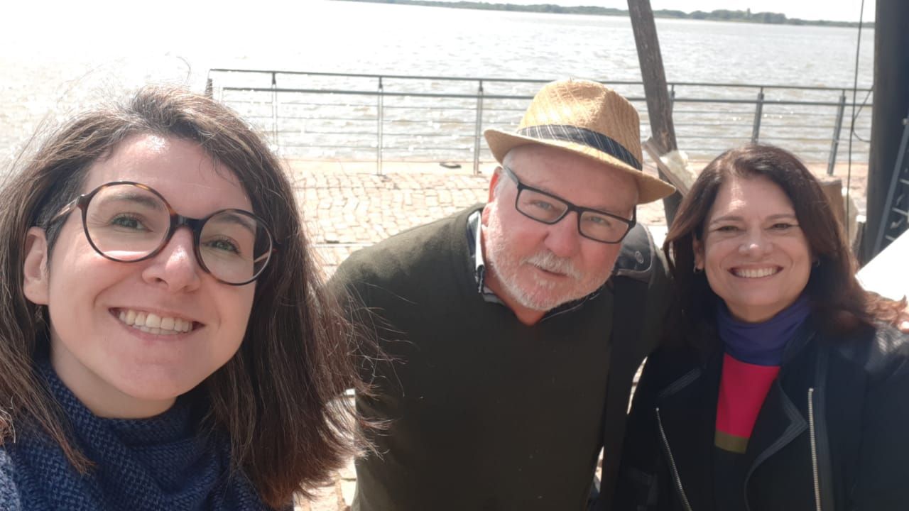 A man at center in a fedora hat and black rimmed glasses looks at the camera, two women stand on either side of him, both with dark hair. Behind them is a waterway and guardrail.