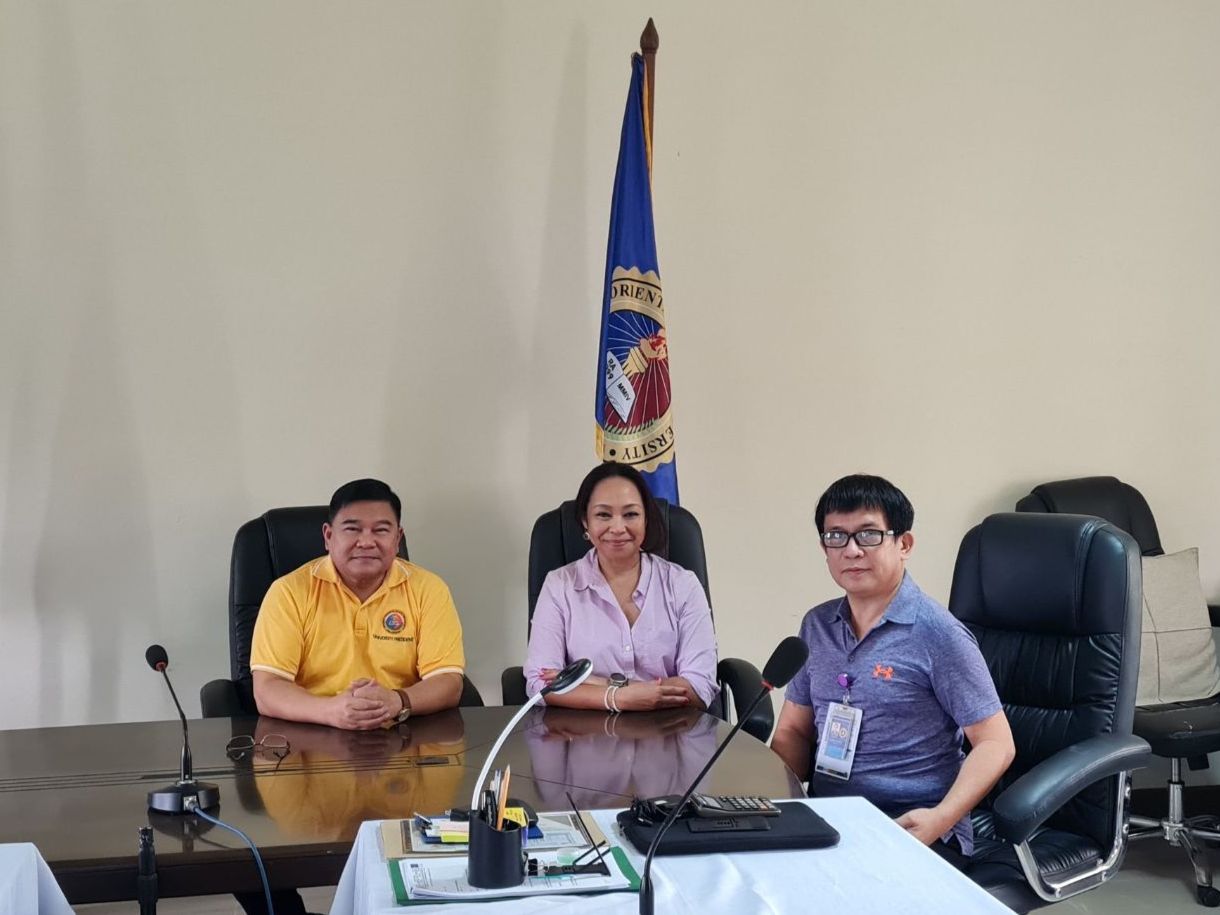 Three people sit at a meeting table with microphones on top of it. A man in a yellow shirt on the left, a woman with a purple button up shirt in the center and a man in a blue shirt on the right. Behind them is a tan wall flanked by two official flags.