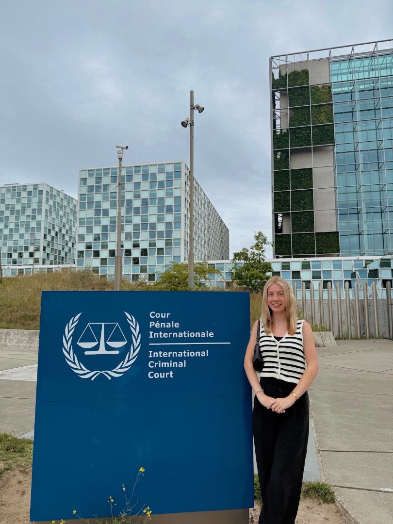 Ashlin wearing a black and white striped top standing by the sign for the International Criminal Court
