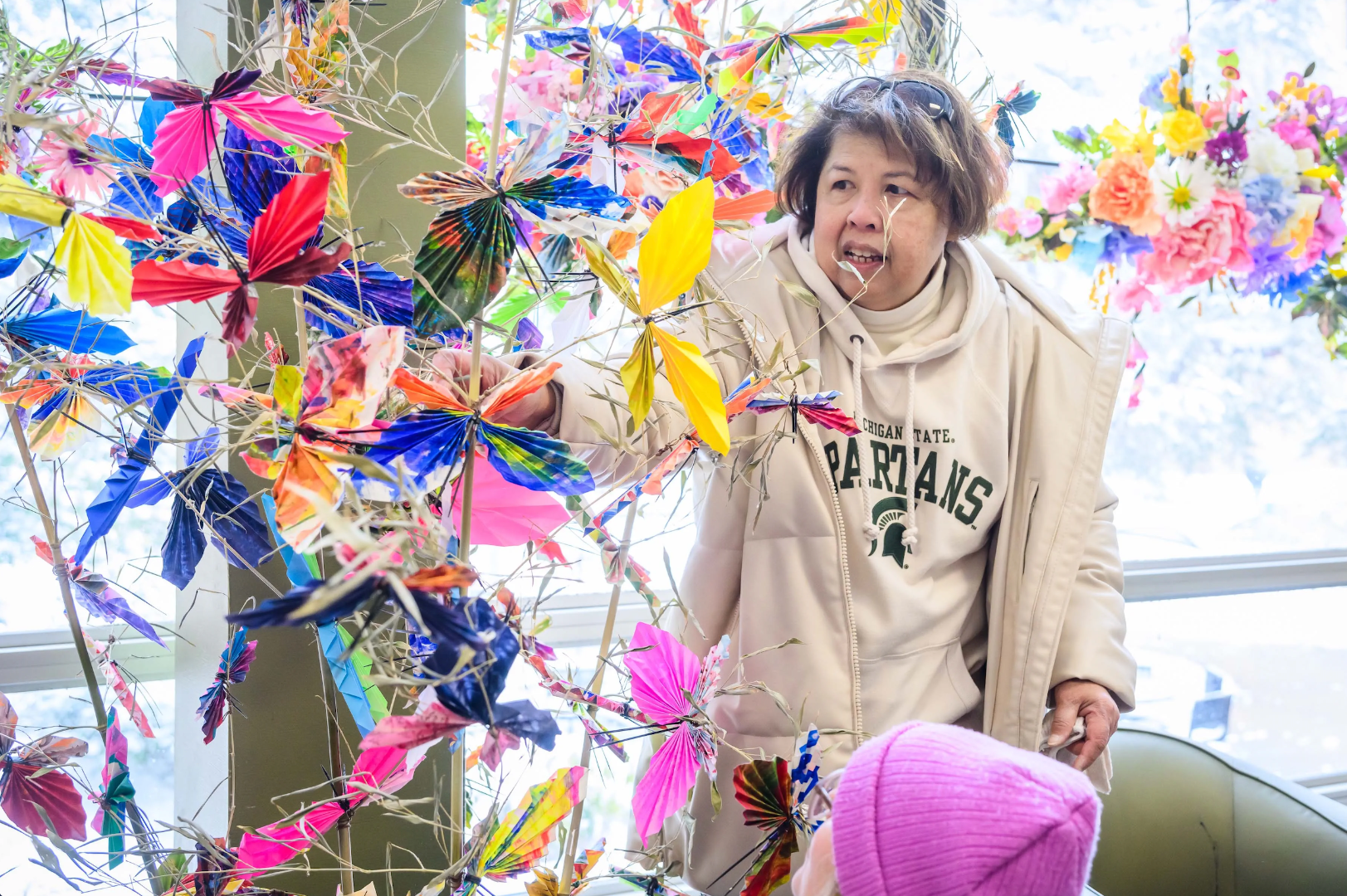 Photo of an older woman wearing a green and white Spartans hoodie tends to a multicolored, multilevel paper butterfly art sculpture.