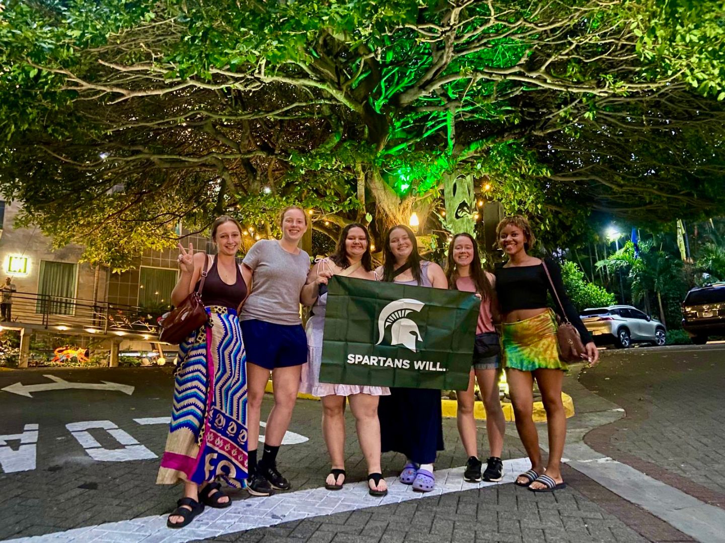 Group of students holding Spartan flag at night under a tree lit with green lights