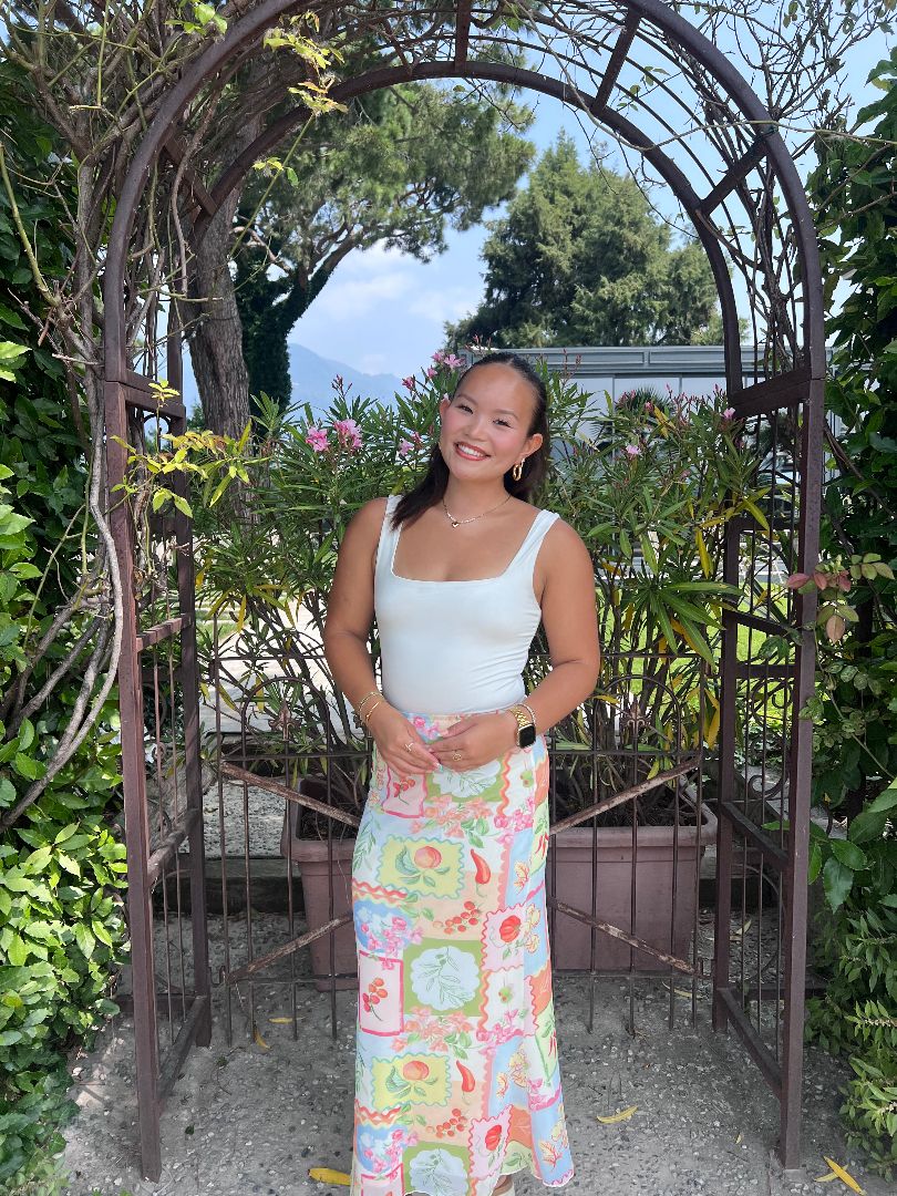 Amelia wearing a white tank top and long floral skirt standing under a garden arch in Italy