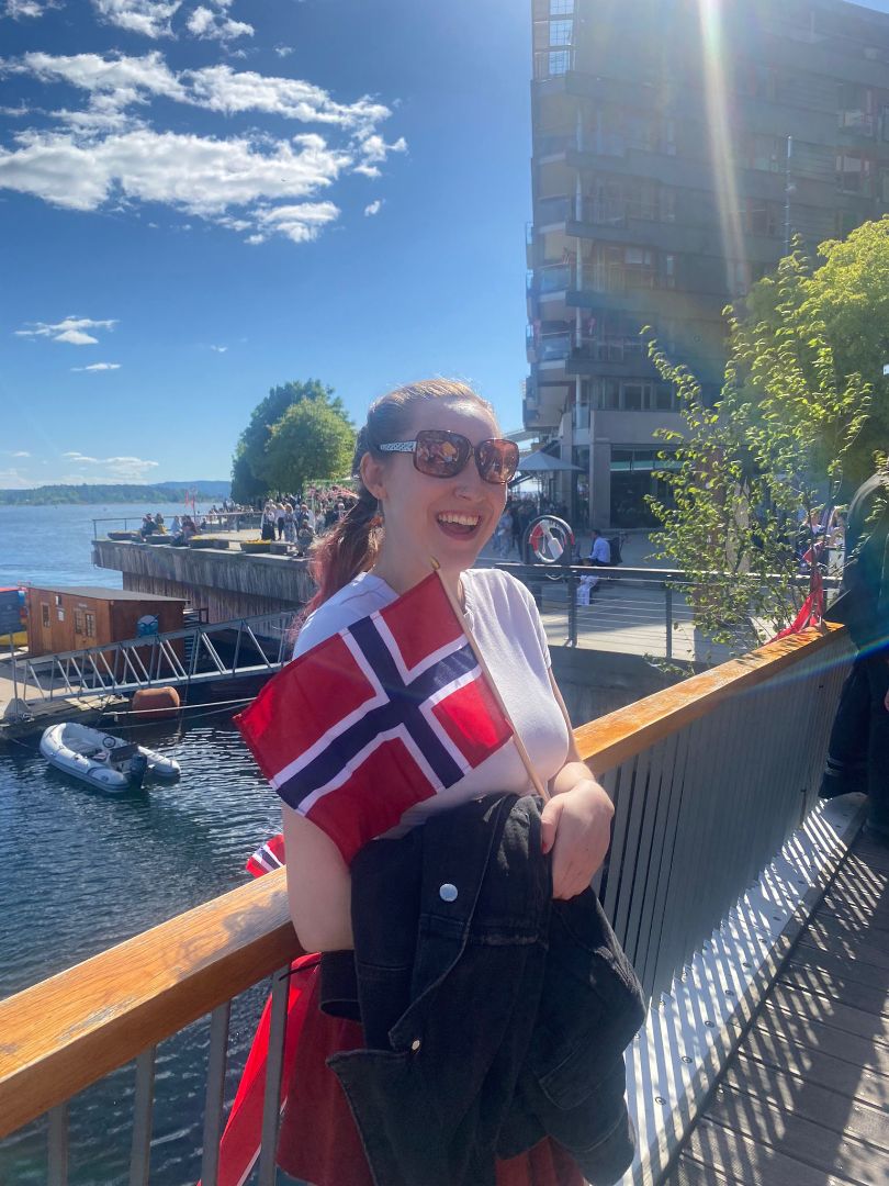 Sophie wearing sunglasses and holding a Norwegian flag posing by a bay in Norway