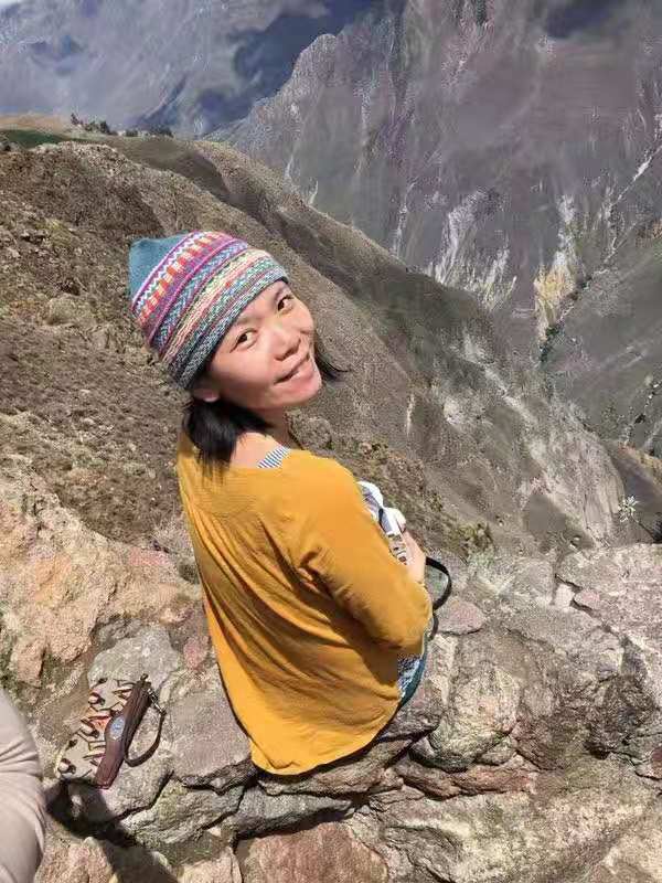 Jing sits on a rocky mountain overlook, smiling and wearing a purple and pink striped skull cap.