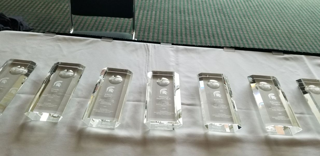 Awards to be presented to recipients are lined up on a table. Seven glass awards with Spartan emblem are pictured.