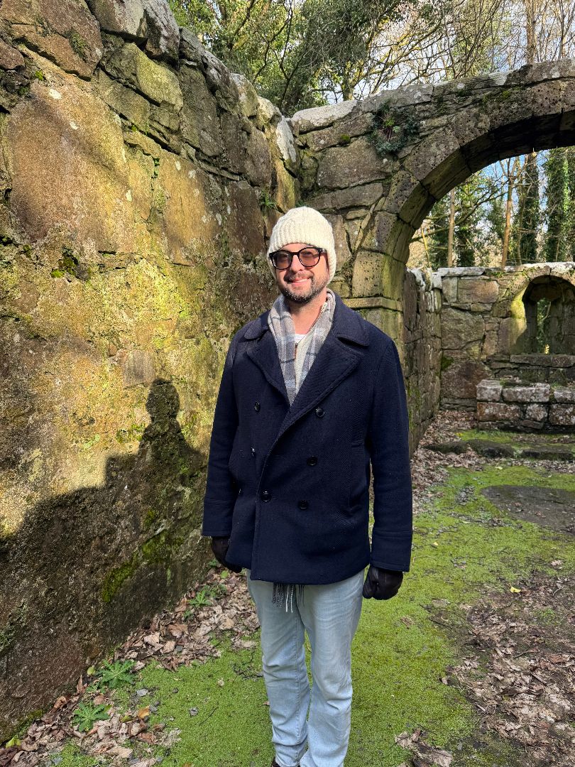Daniel wearing a white ski hat and navy coat standing underneath an ancient stone arch in Ireland