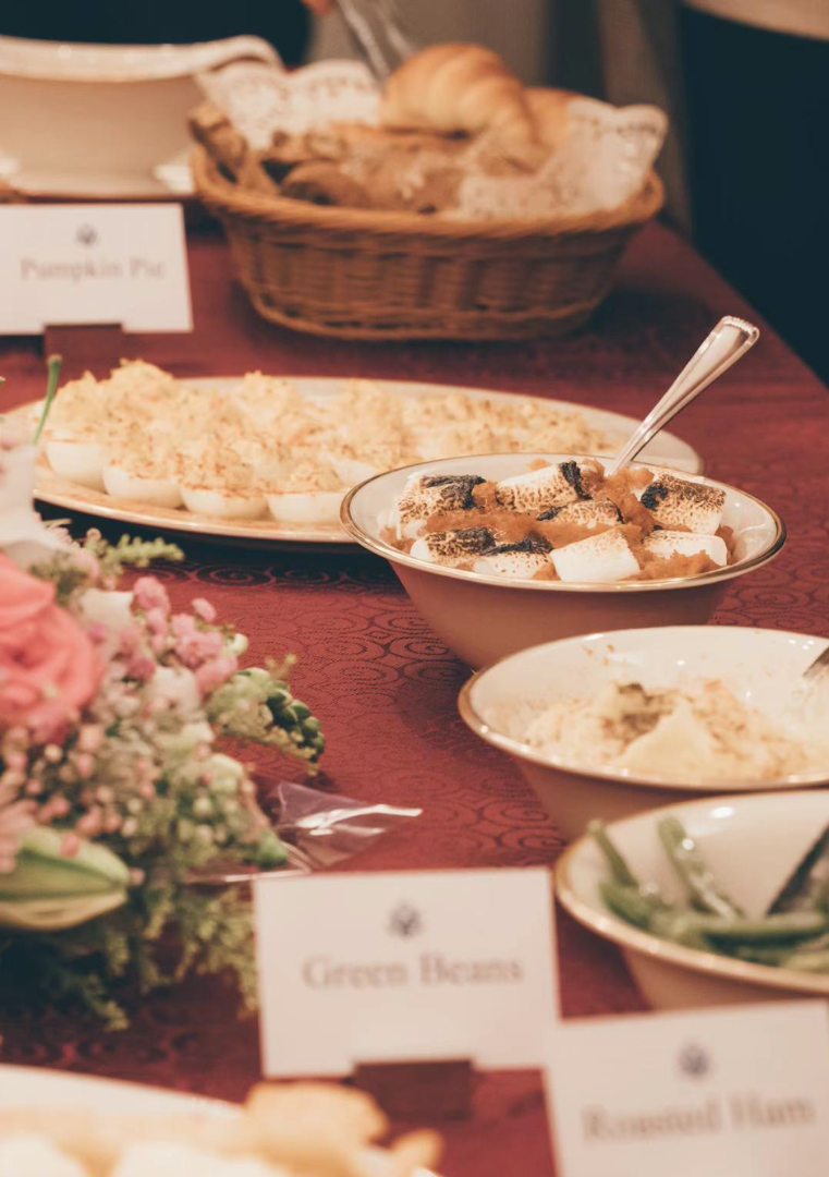 Close up image of a red table cloth with flowers and bowls of food: sweet potatoes with toasted marshmallows, mashed potatoes, and deviled eggs.