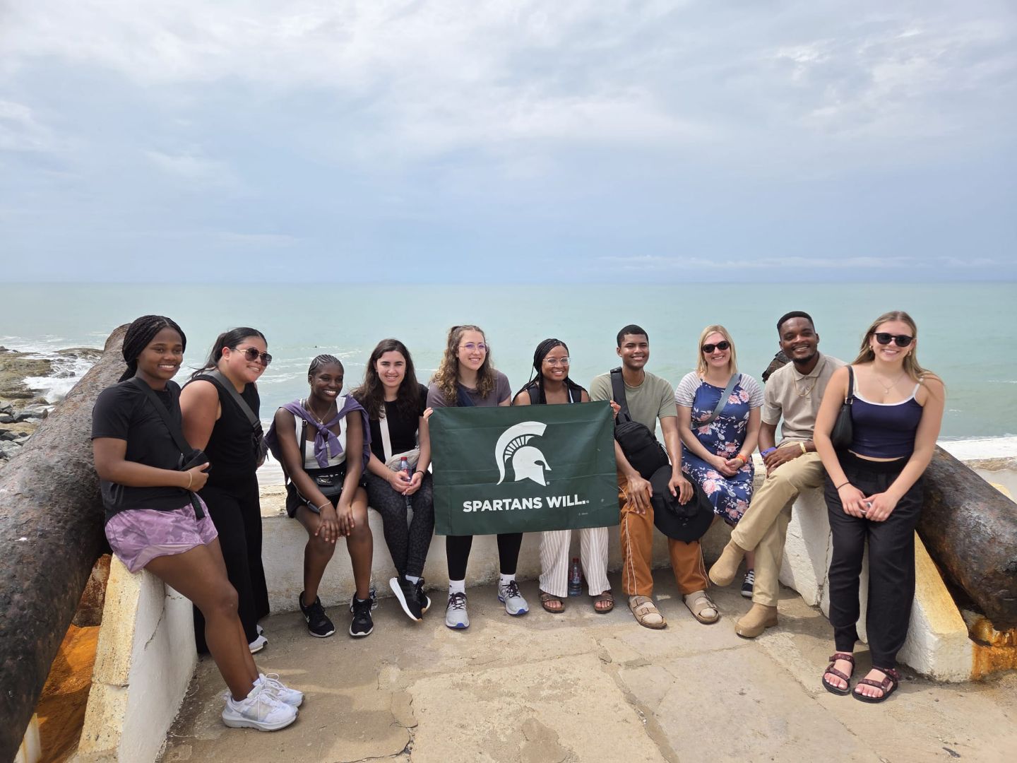 Group of students holding the Spartan flag on a stone balcony overlooking the ocean