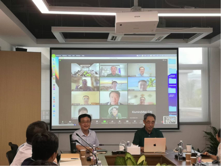 Hu Feng and Qi sit at the head of a conference table, smiling with virtual participants pictured on a screen behind them.