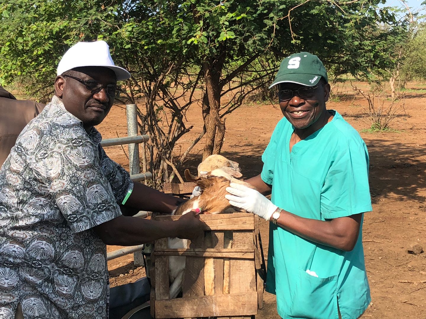 John B Kaneene stands with a colleague in an arid landscape holding a goat.