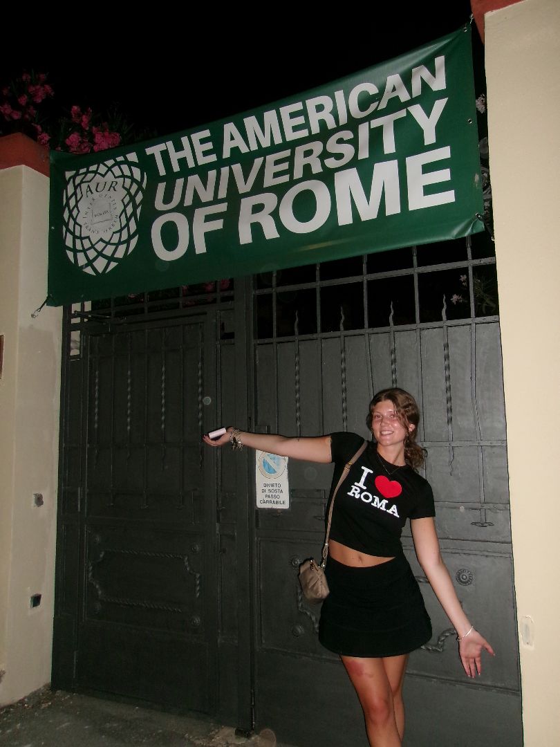 Elizabeth wearing an I love Roma tee shirt standing in front of a door with a banner that reads The American Universtiy of Rome