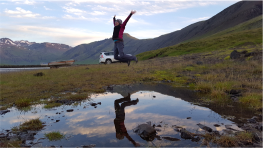 A person jumps high above a small body of water, arms outstretched and legs bent underneath them, their reflection in the water below. There is a mountain range in the background and the landscape is dotted with green plant life. 