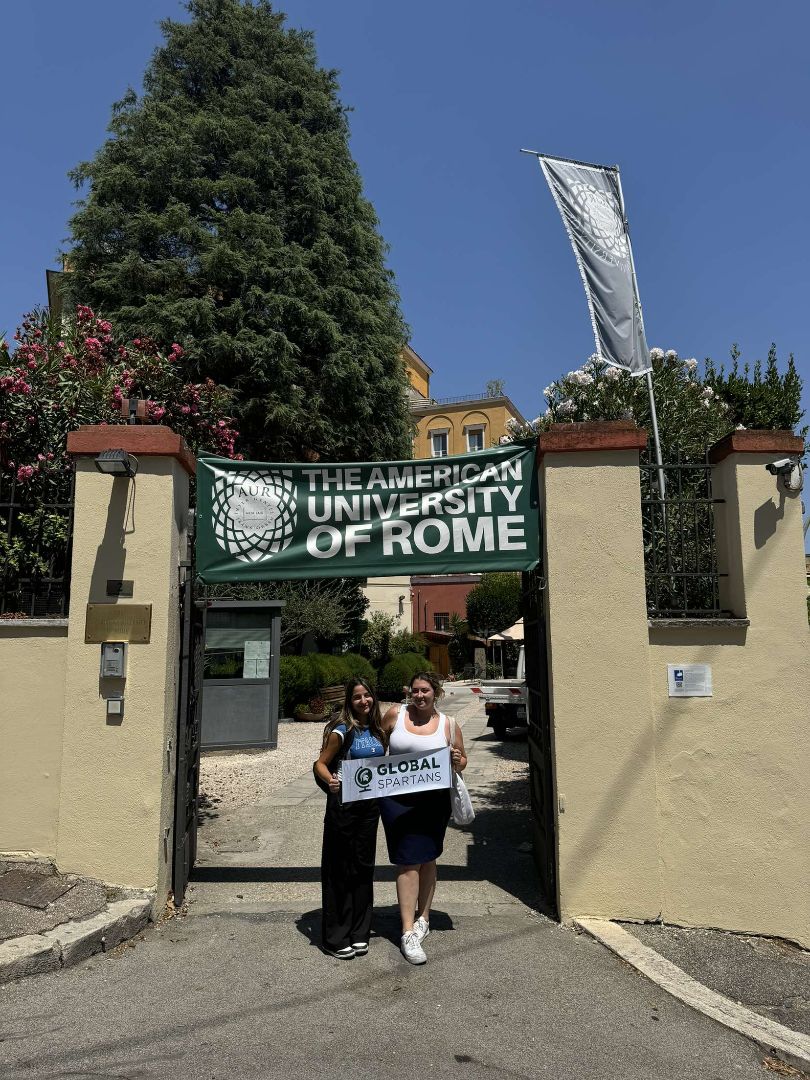 Christina and friend holding a Global Spartans banner in front of the American University of Rome entrance