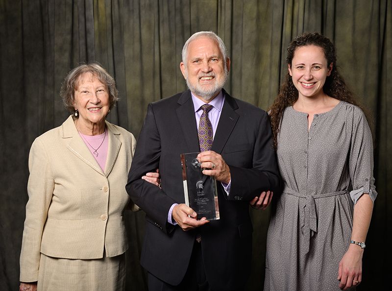 John Metzler [male] pictured  holding award with two female guests- one female on each side. John is in the center holding award.