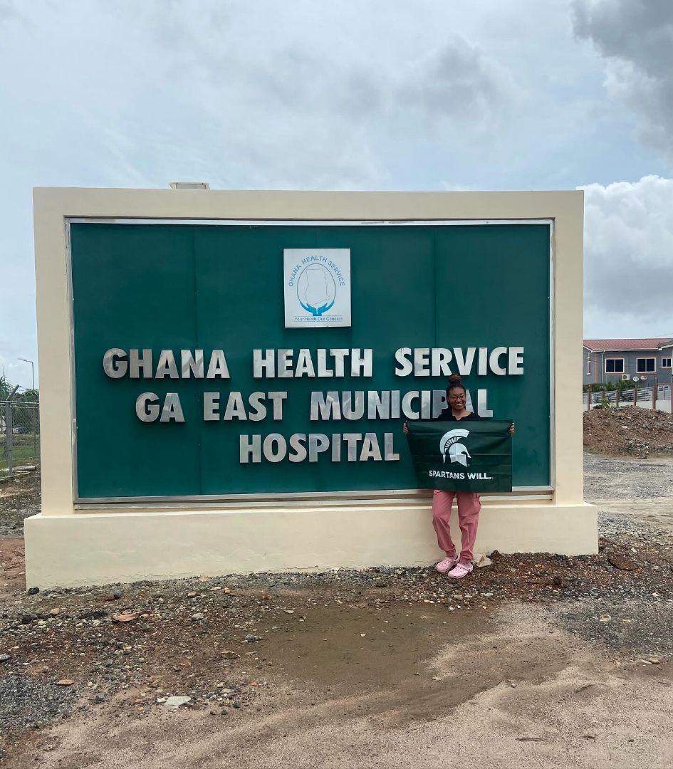 Danielle holding Spartan flag in front of sign that reads Ghana Health Service