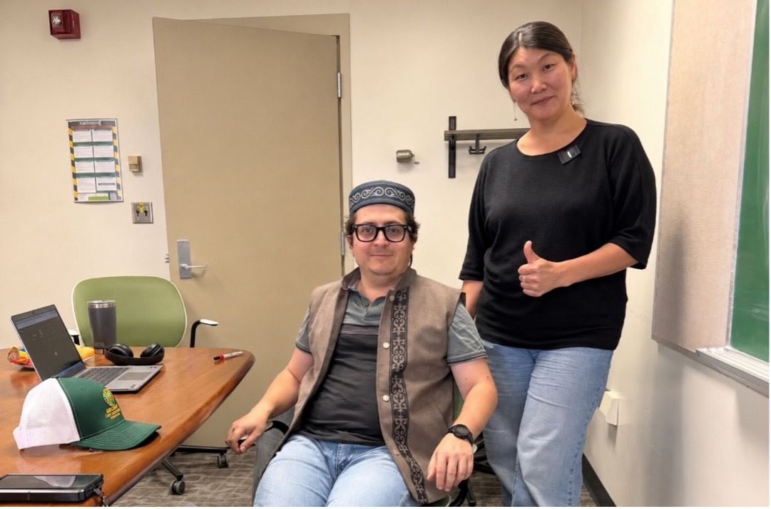 Dukenbayeva and Hoyos-Villegas in a university classroom, with one seated at a desk wearing a traditional patterned hat and vest while the other stands beside them giving a thumbs-up.